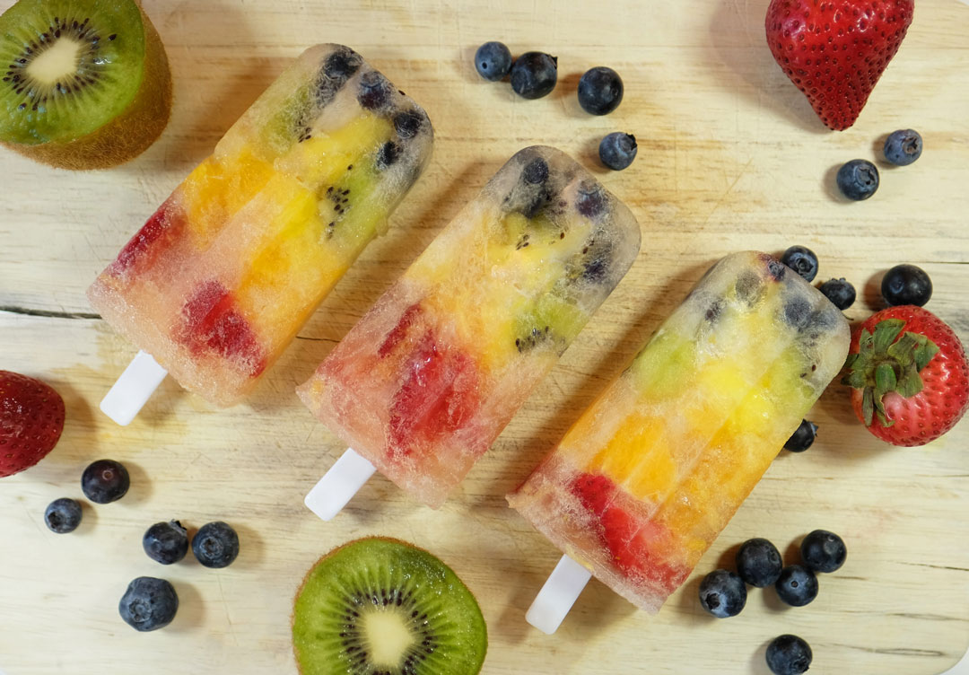Rainbow popsicles on a cutting board with some fruit